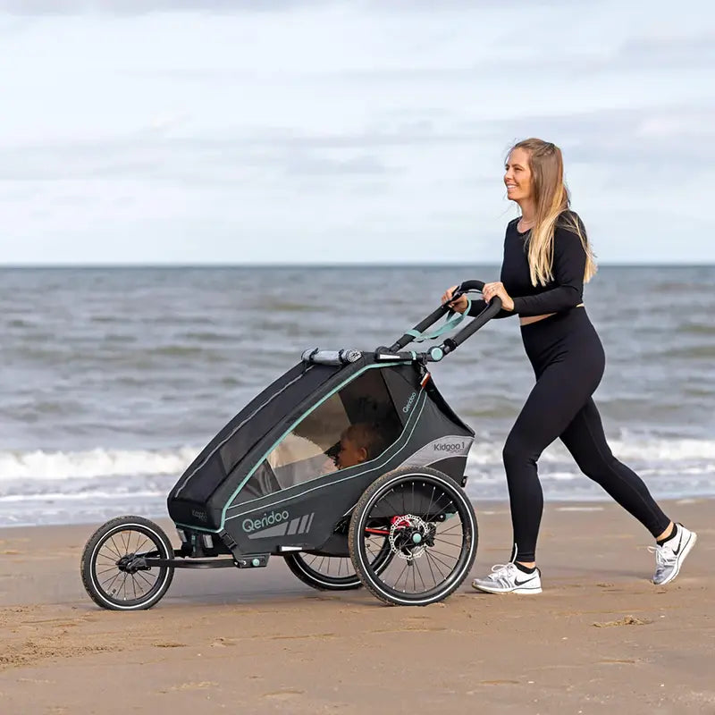 Frau schiebt Qeridoo Kidgoo 1 Fahrradanhänger mit Joggerrad als Jogger am Strand