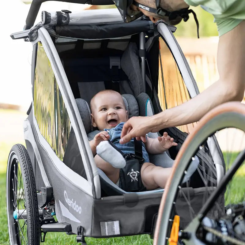 Lachendes Baby im grauen Qeridoo Fahrradanhänger, angeschnallt mit Gurt, neben einem Fahrrad auf der Wiese