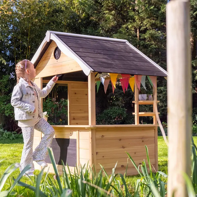 Mädchen spielt an einem Holzspielhaus mit Theke, dunklem Dach, Tafel und bunter Wimpelkette im Garten